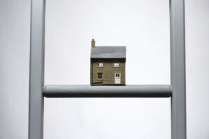 A miniature house perched atop a ladder against a plain background.
