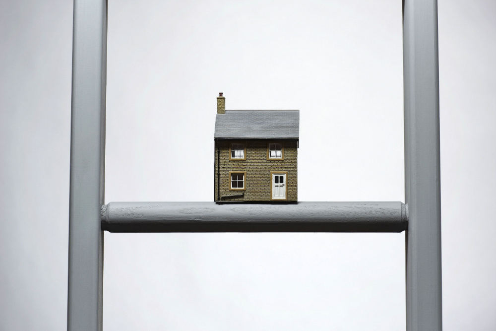 A miniature house perched atop a ladder against a plain background.