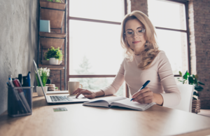 A woman sits at a desk, working on a laptop with a pen in hand.